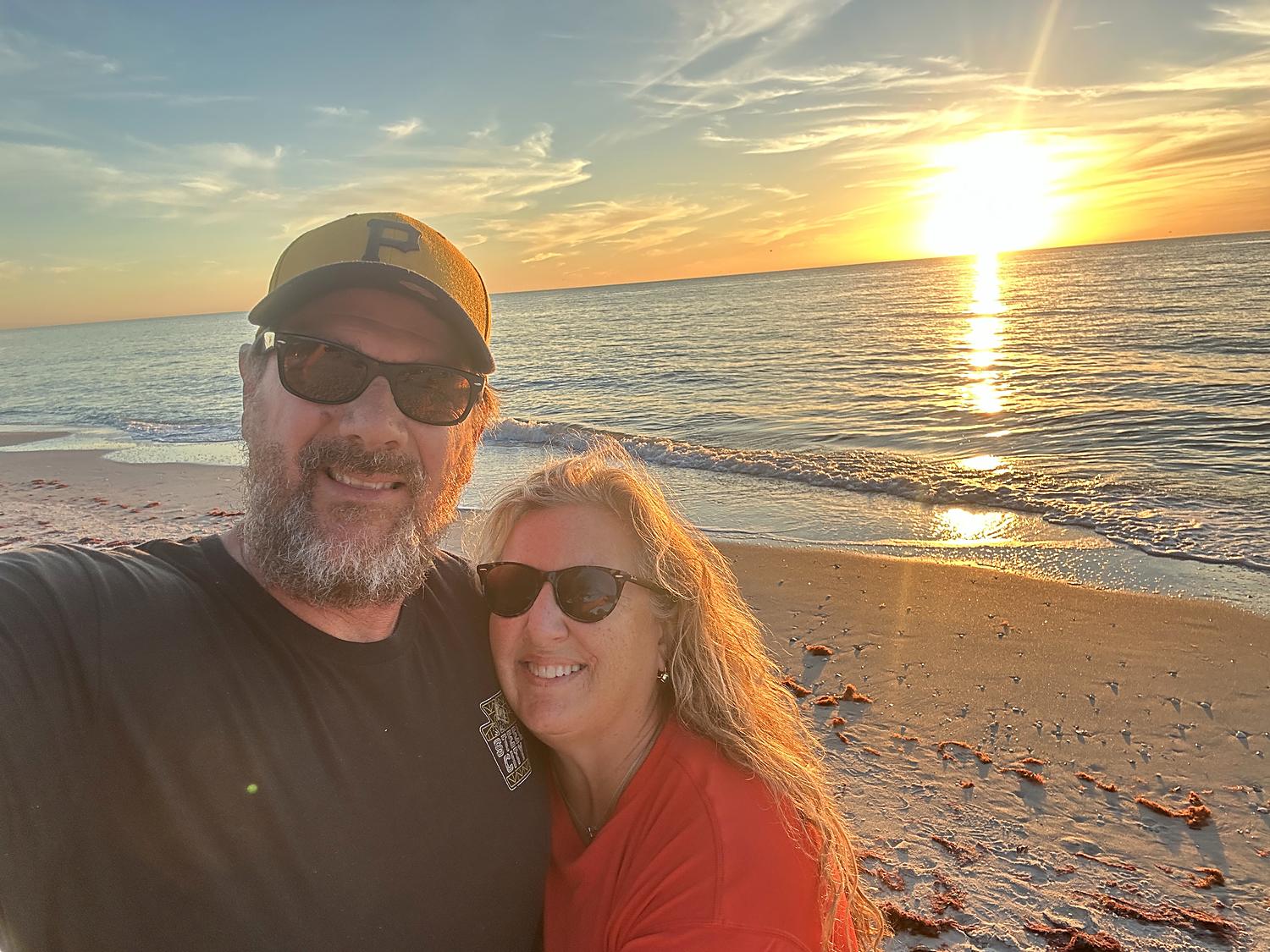 John and Jacque on the beach at sunset, the sun low over the water behind them.