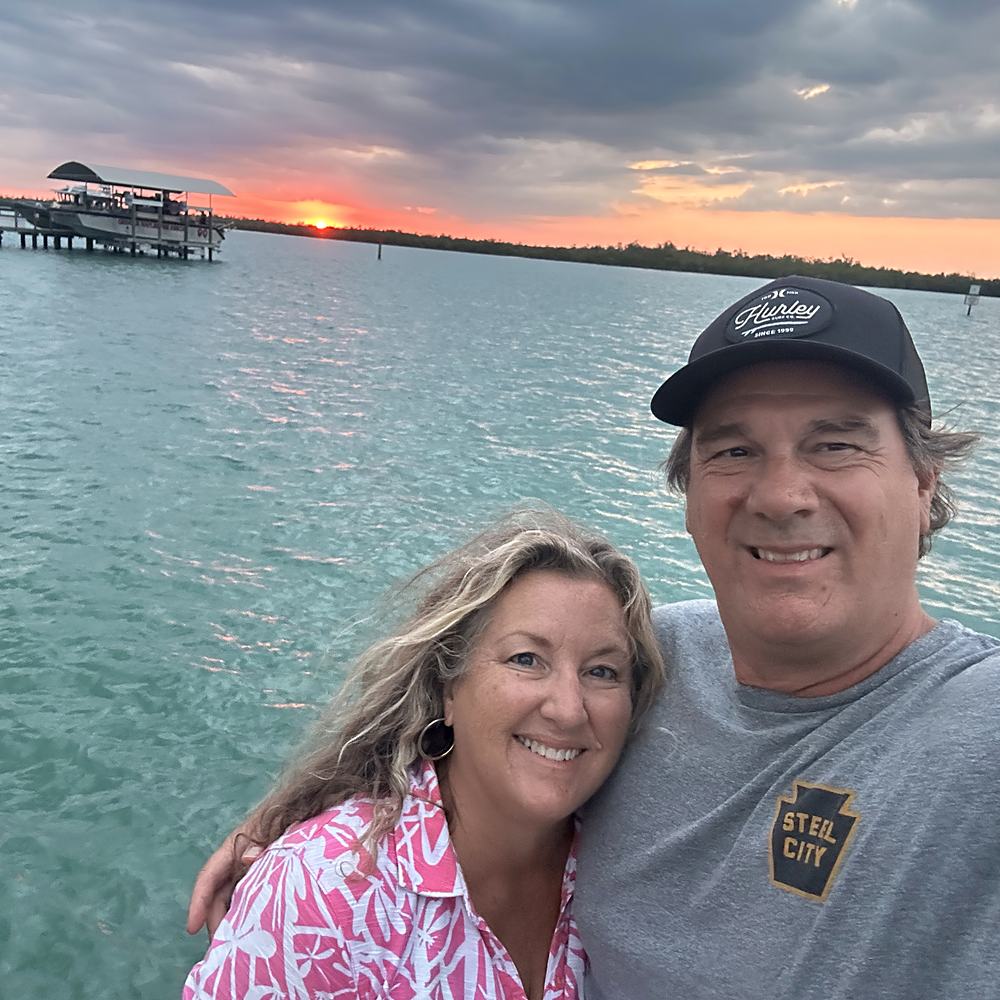 John and Jacque at the water's edge as the sun goes down behind a pier.